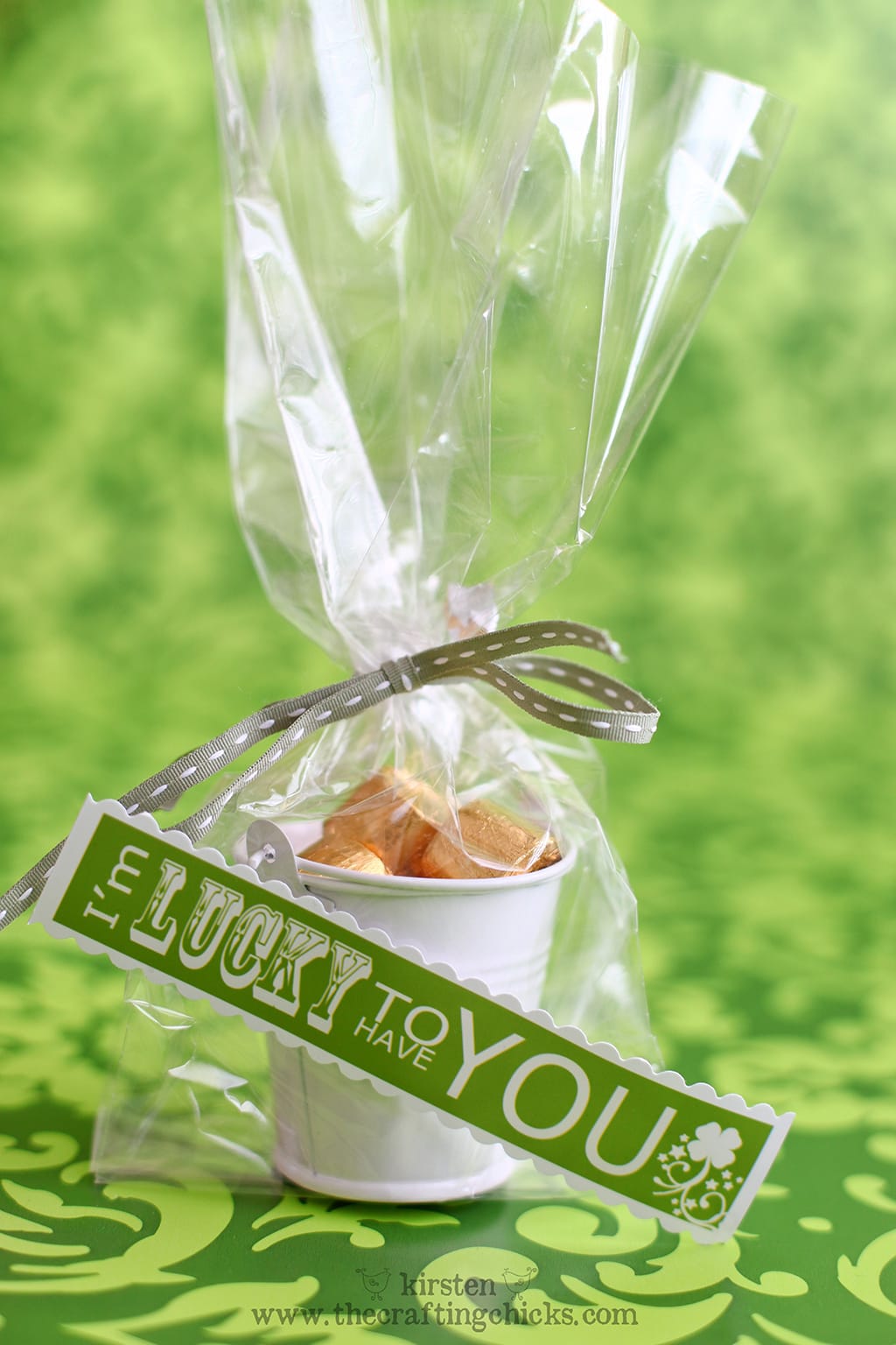 Green background withe small white buckets filled with gold wrapped candies in a clear cellophane bag and a green ribbon. A printable tag is attached that says "I'm lucky to have you."
