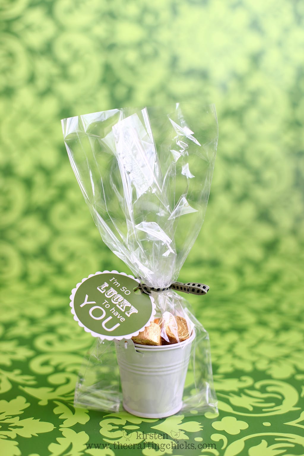 Green background withe small white buckets filled with gold wrapped candies in a clear cellophane bag and a green ribbon. A printable tag is attached that says "I'm so lucky to have you."