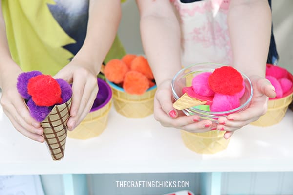 Children's hand holding a cardboard ice cream cone filled with pompoms and a glass bowel filled with pompoms for ice cream