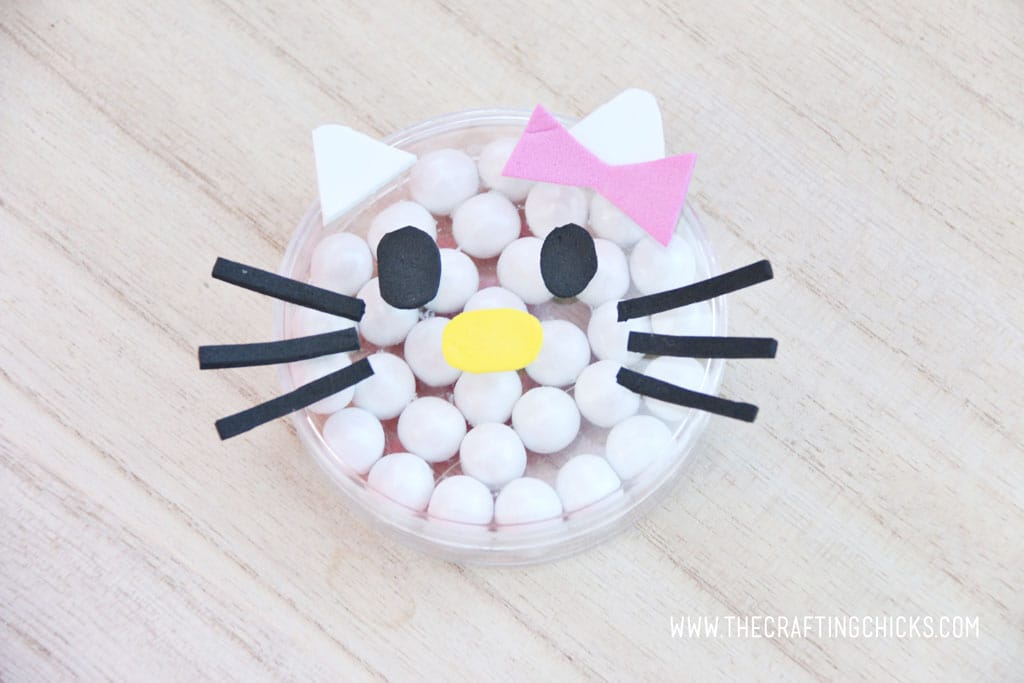 Pink foam bow added to a clear round favor container filled with white Sixlets to make a Hello Kitty Face