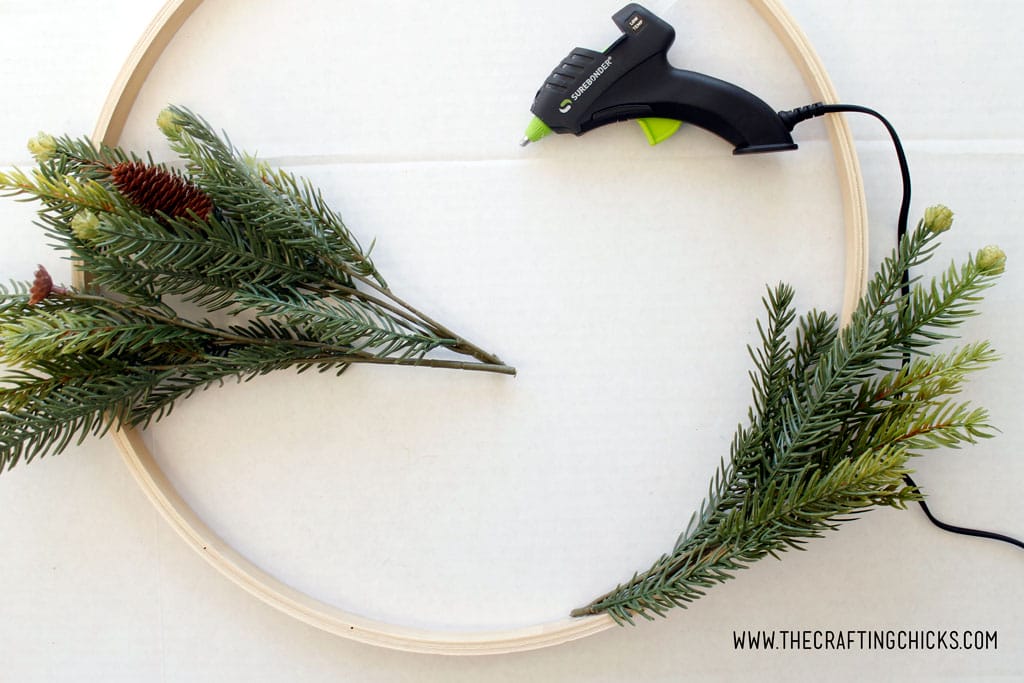 Using a hot glue gun to attach faux pine branches to an embroidery hoop to make a hoop wreath.