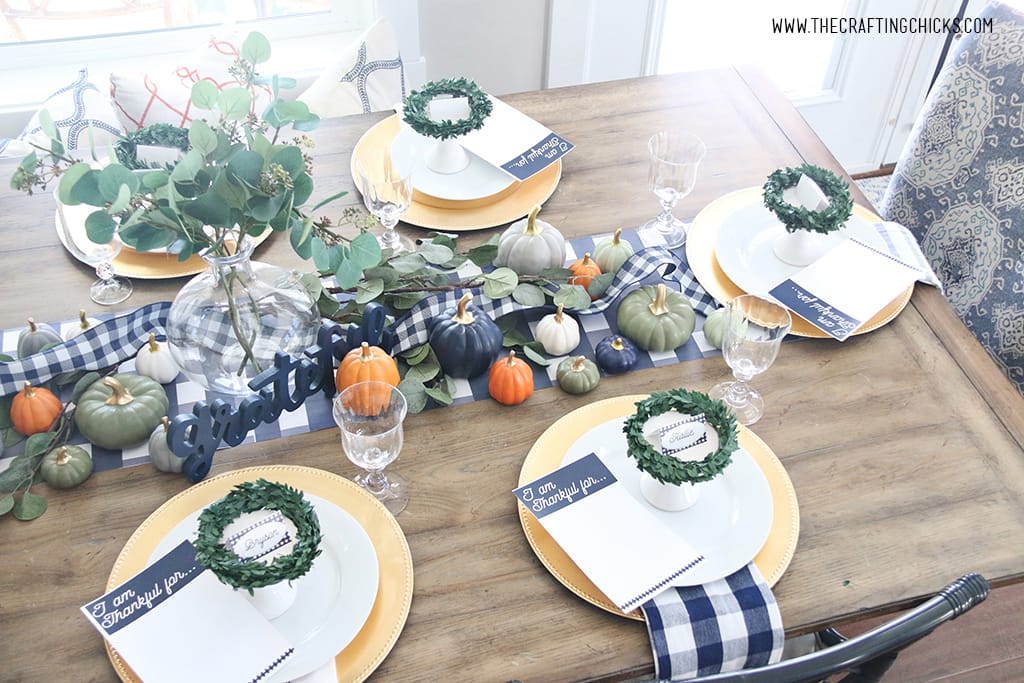 Styled Thanksgiving table with gold chargers and white plates. Navy and white check print napkins and table runner. With mini pumpkins and small wreaths to decorate.