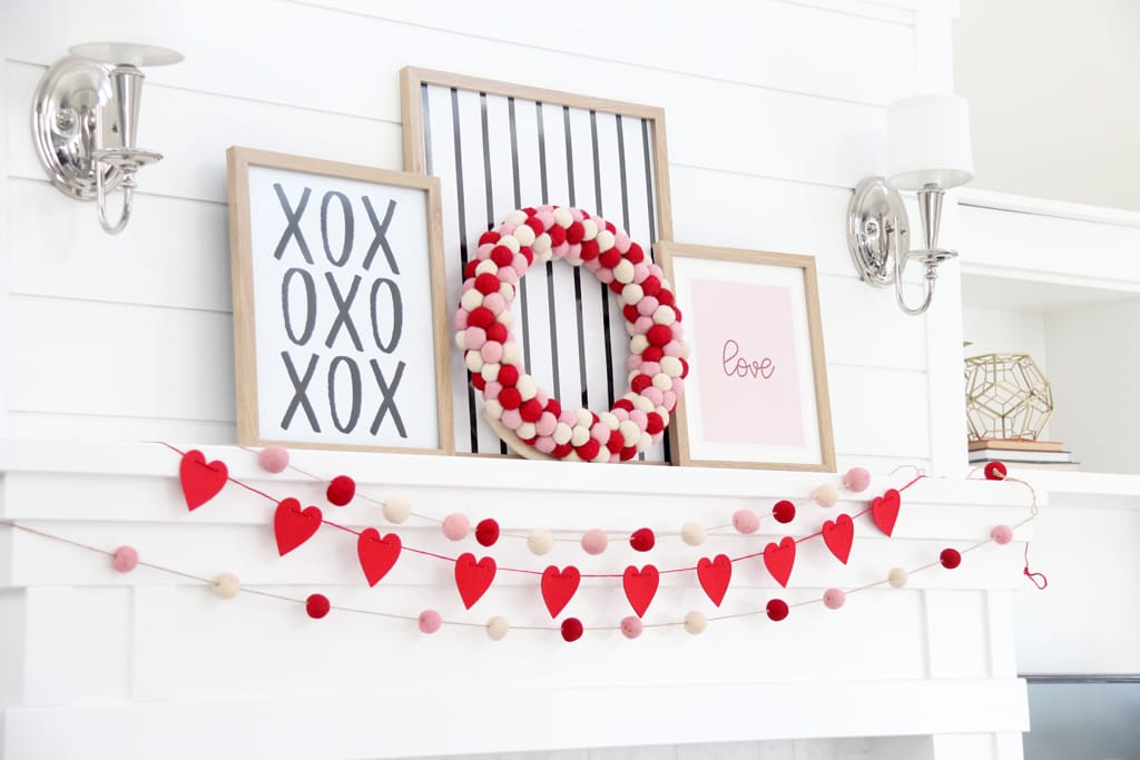 White mantle with red heart banner and pink, red, and white pom pom banner. Three pictures in frames one with XOXO in black and white, one with black and white stripes, and one with Pink background and the word "Love" in red.