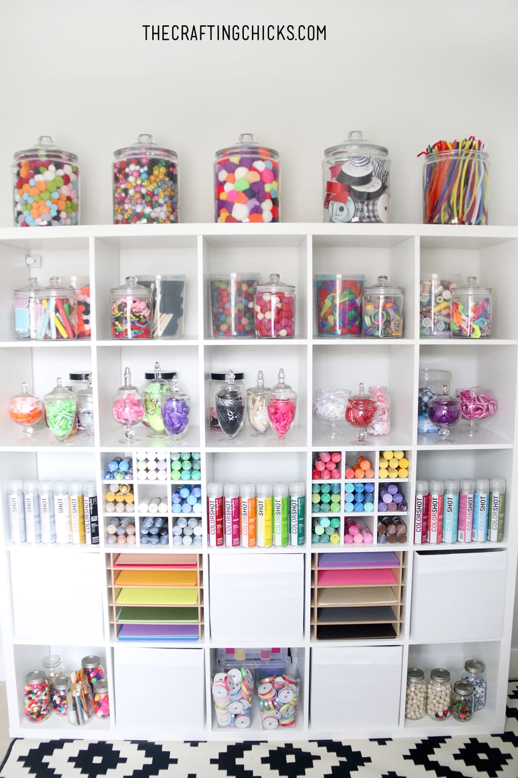 White shelves decorated with colorful jars filled with craft supplies, colorful paper, and colorful paint.