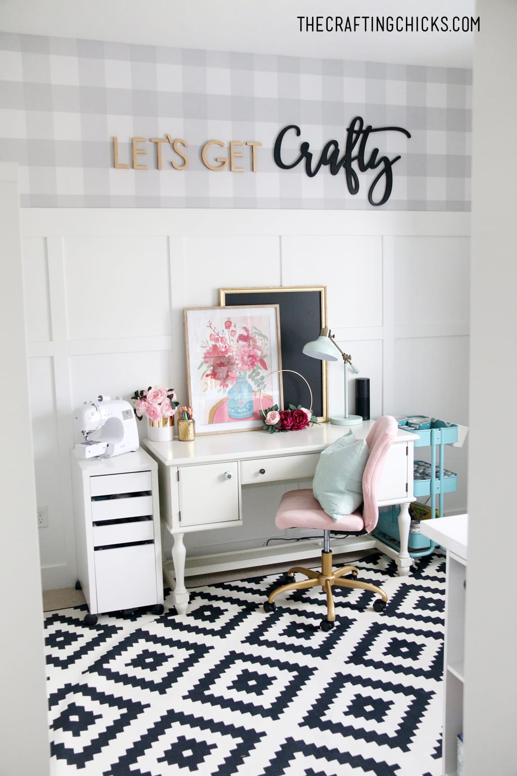 White desk with pink plush chair and sewing machine on the side. Gray and white plaid wallpaper with Let's Get Crafty wood cutout letters.