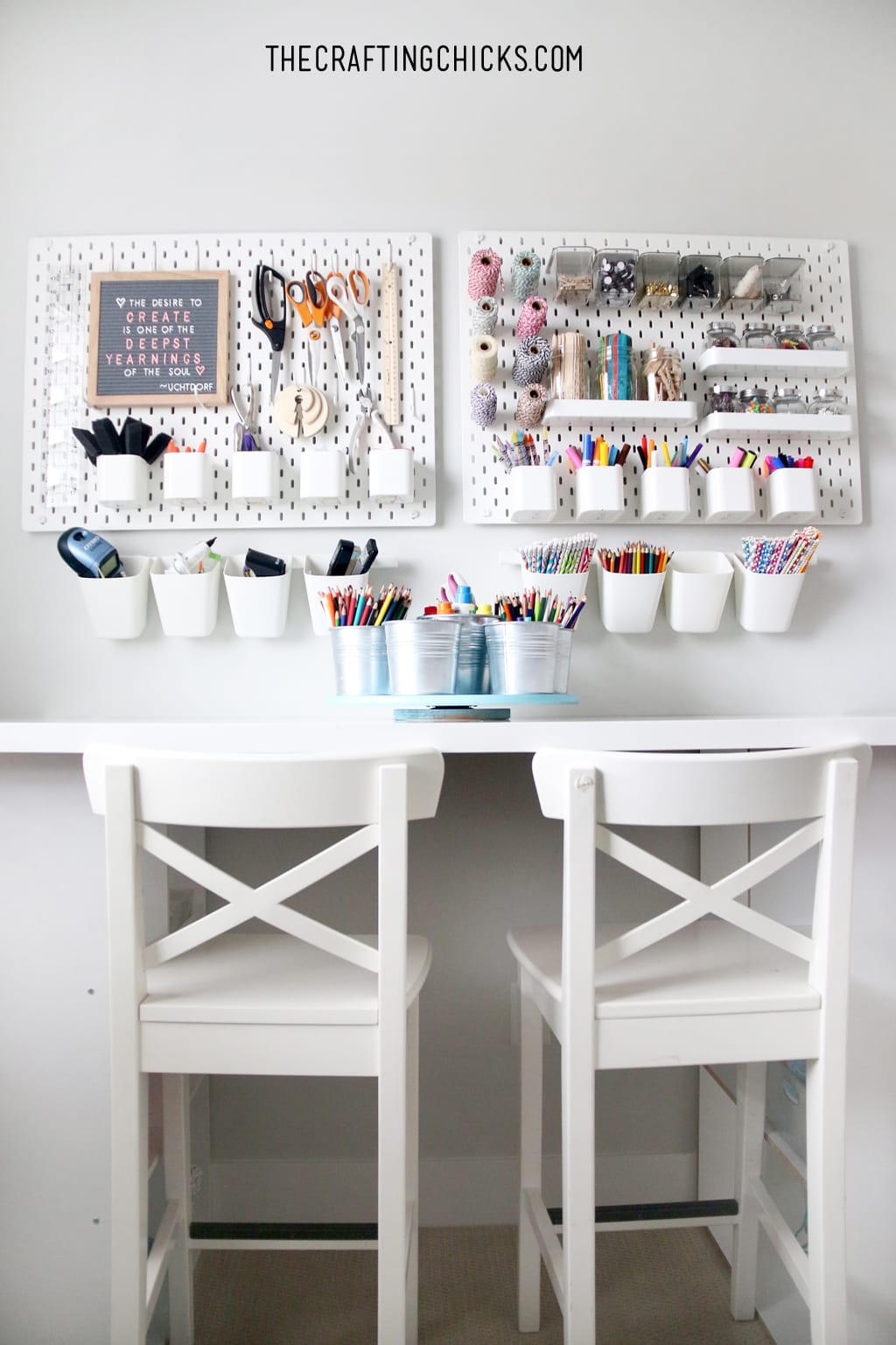 Wall with white peg board and craft supplies on it. Table and white stools in front for a crafting space.