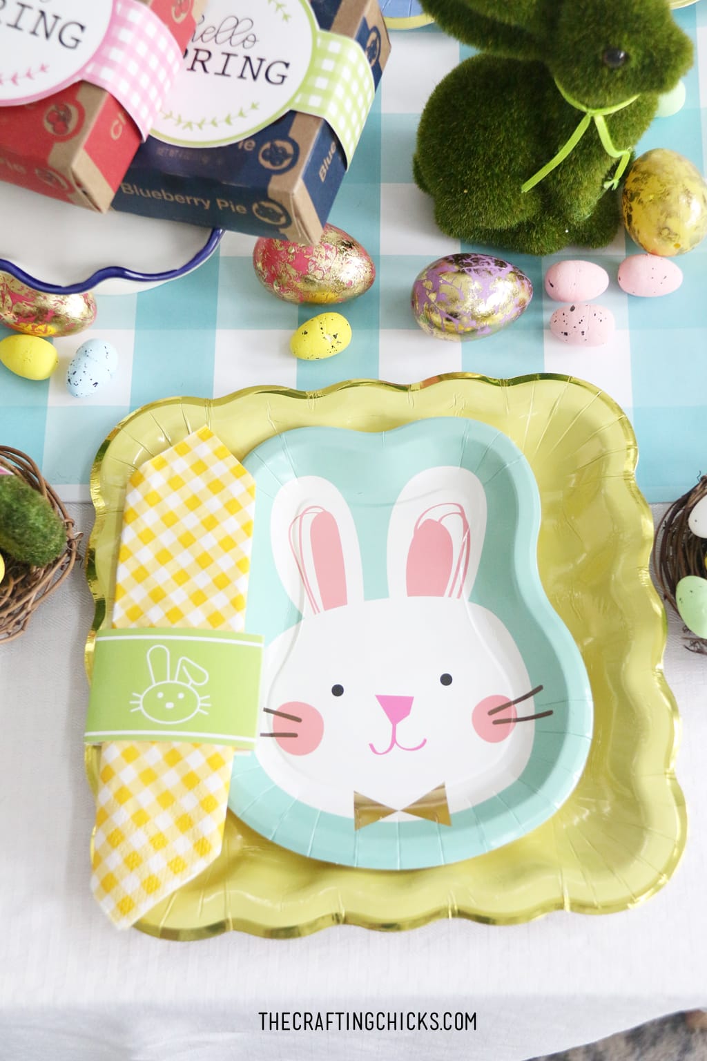 Easter table for kids with bunny paper plates, yellow gingham napkin and bunny decorations. Big Pink bunny printable backdrop