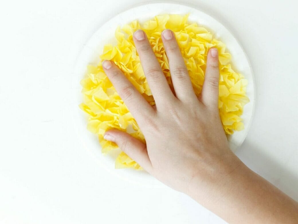 White paper plate filled with yellow tissue paper, with hand pressing down on it.