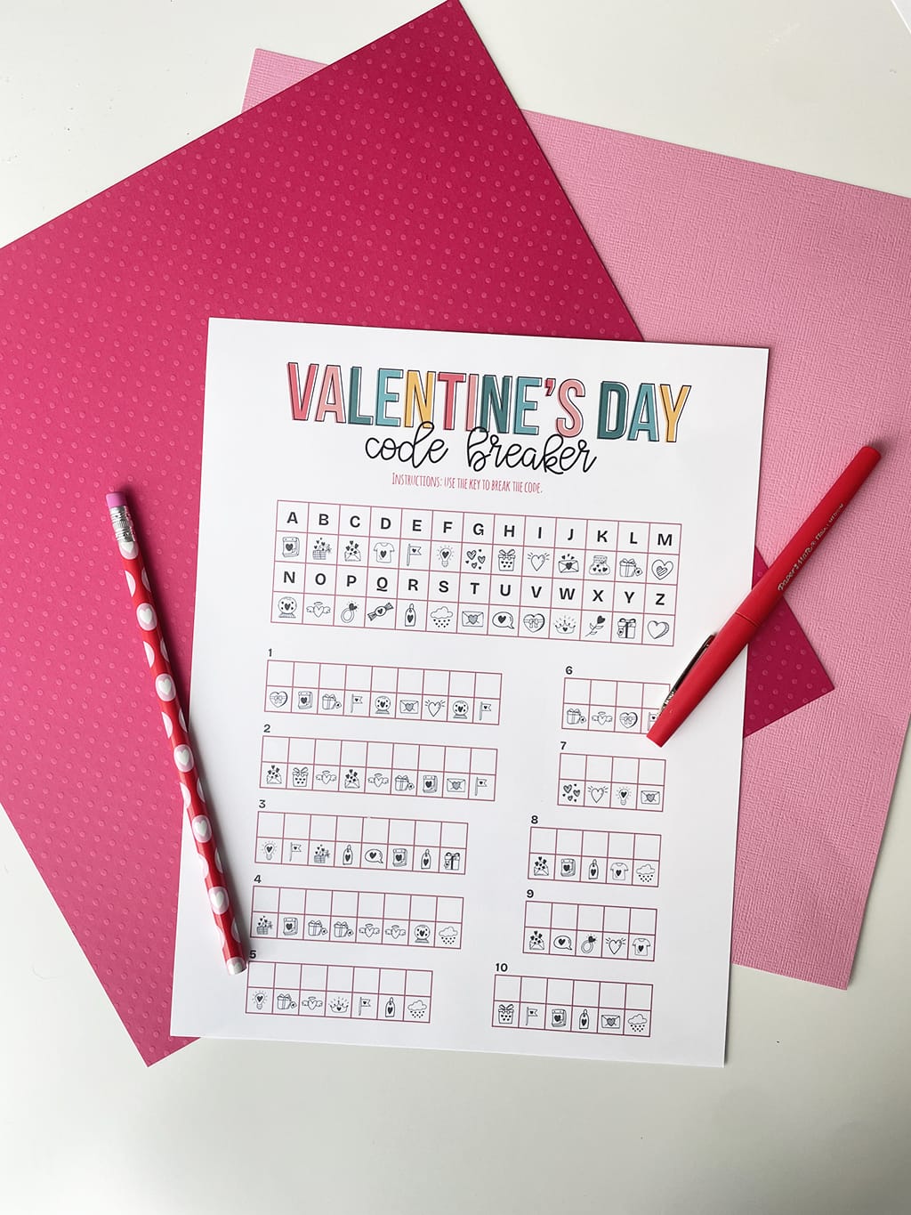 Valentines Day Code Breakers Printable Game printed out and placed on a dark pink and light pink paper backdrop. With a red fineline marker and pencil.