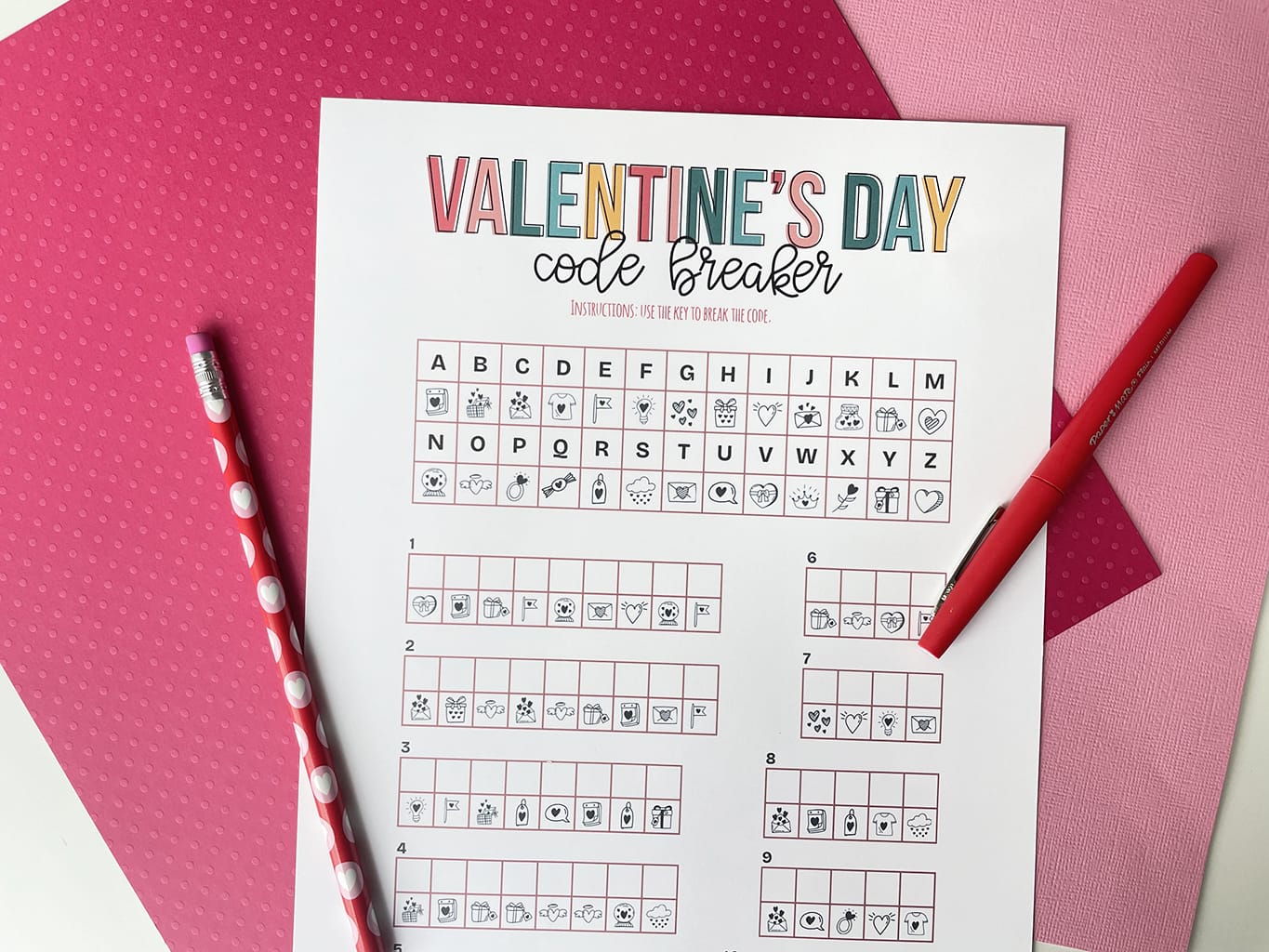Printable of the Valentine's Day Code Breaker printed out and placed on a white table, with dark pink and light pink paper behind it. Red fine line marker on right and a valentine pencil on the left.