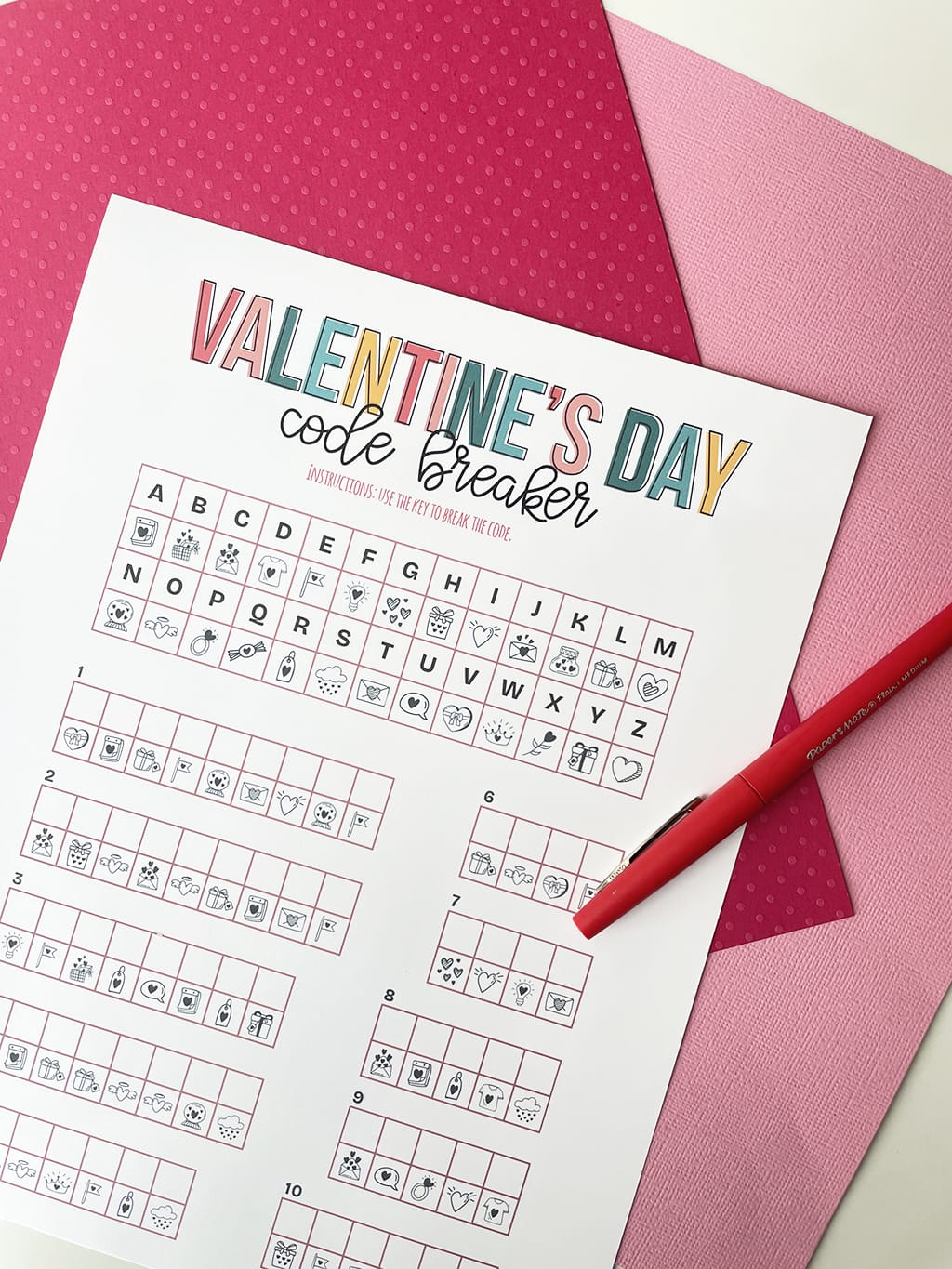 Printable of the Valentine's Day Code Breaker printed out and placed on a white table, with dark pink and light pink paper behind it. Red fine line marker on right and a valentine pencil on the left.