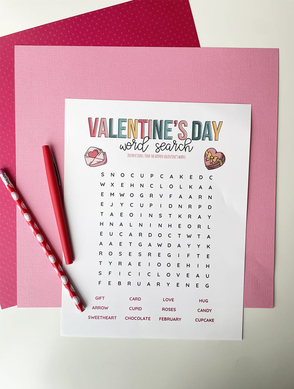 Valentines Day Word Search Printable Game printed out and placed on a dark pink and light pink paper backdrop. With a red fineline marker and valentine pencil.