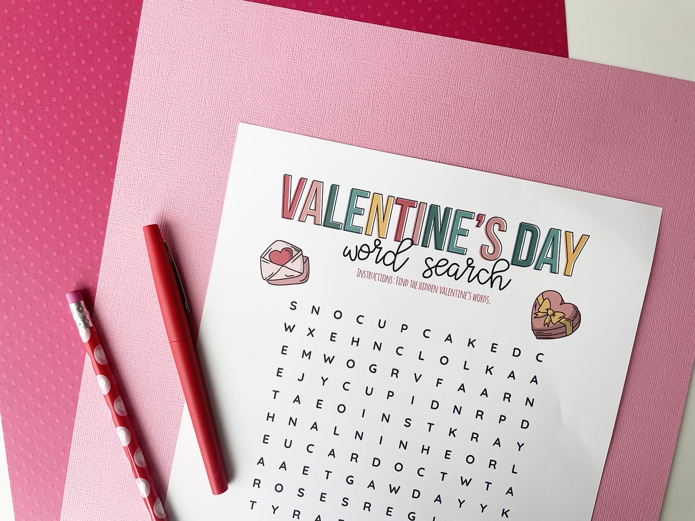 Valentine's Day Word Search Printable, printed out and placed on a light pink paper and a dark pink paper background. Valentine pencil and red fine line marker on the side.