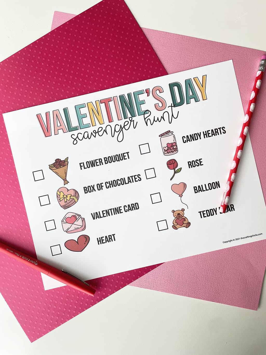 Printable of the Valentine's Day Scavenger Hunt printed out and placed on a white table, with dark pink and light pink paper behind it. Red fine line marker is on the left side and a valentine pencil is on the right side.