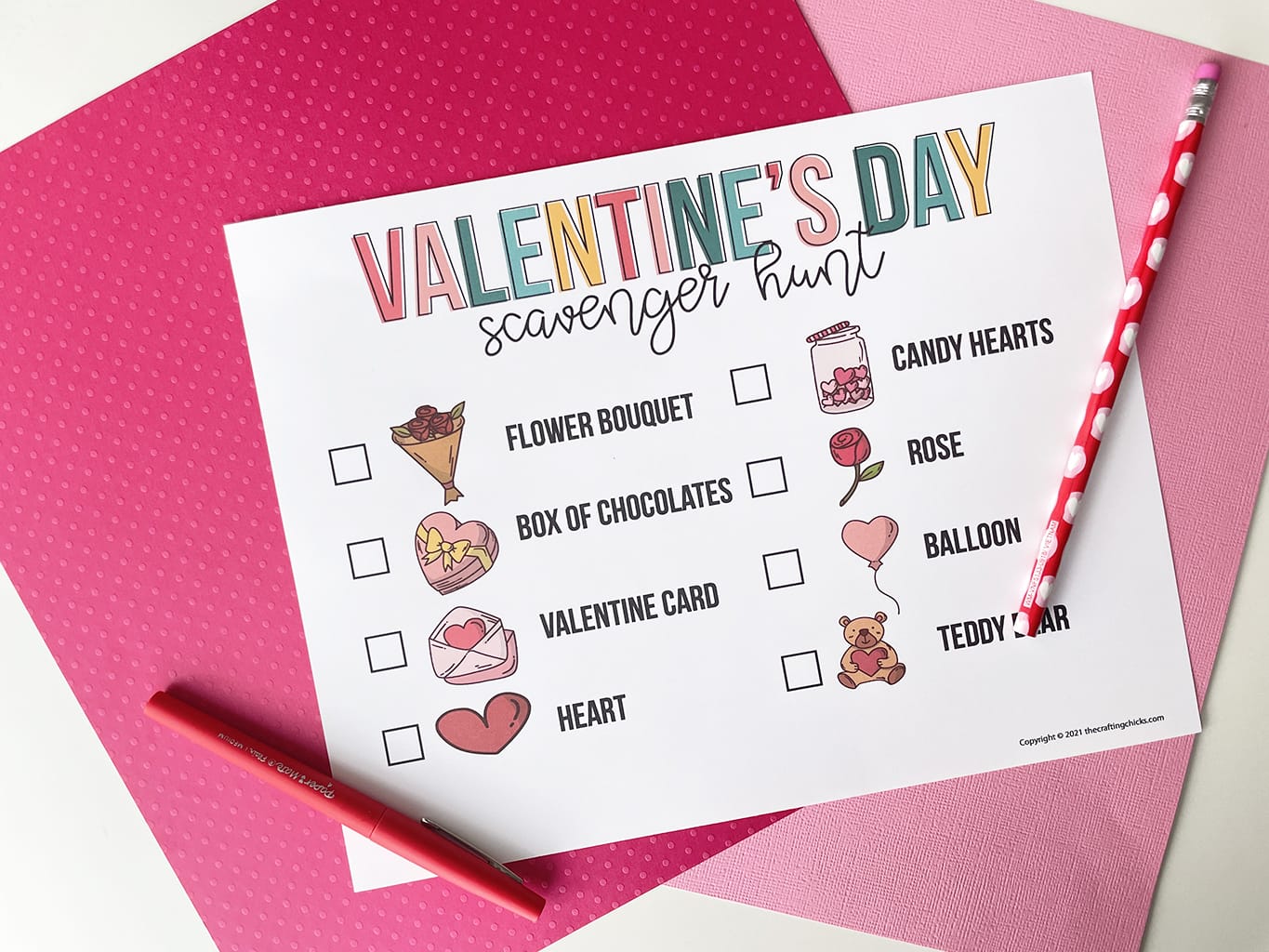 Printable of the Valentine's Day Scavenger Hunt printed out and placed on a white table, with dark pink and light pink paper behind it. Red fine line marker is on the left side and a valentine pencil is on the right side.