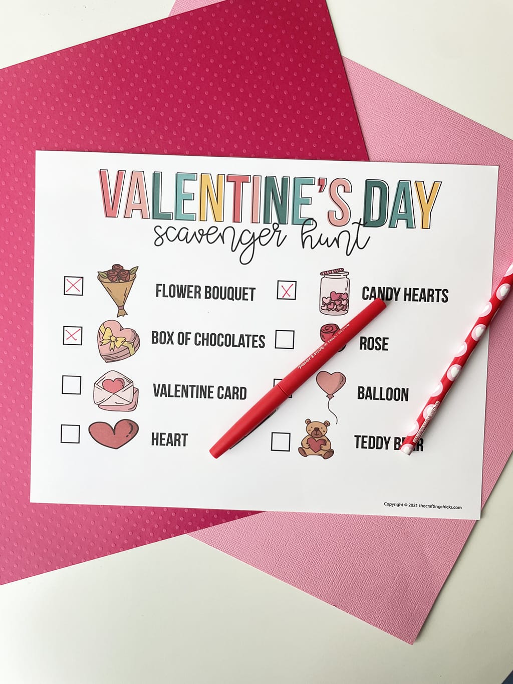 Valentines Day Scavenger Hunt Printable Game printed out and placed on a dark pink and light pink paper backdrop. With a red fineline marker that has written in some of the answers.