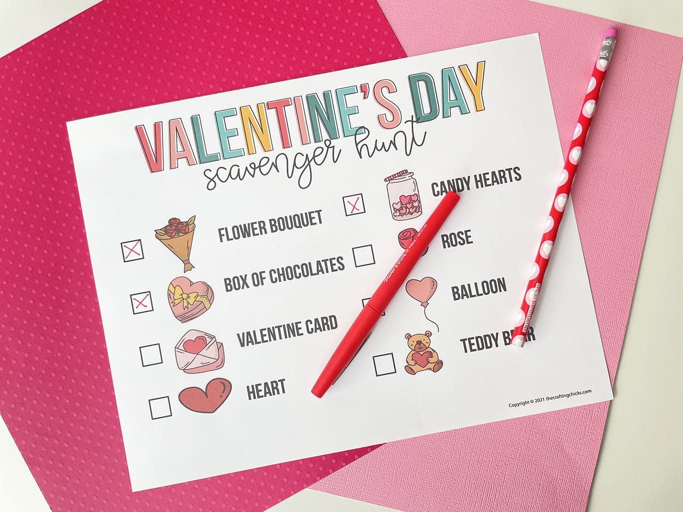 Printable of the Valentine's Day Scavenger Hunt printed out and placed on a white table, with dark pink and light pink paper behind it. Red fine line marker has crossed off some of the boxes.