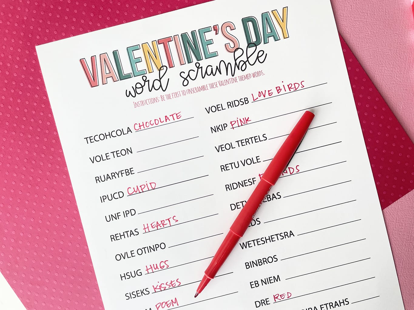 Valentines Day Word Scramble Printable Game printed out and placed on a dark pink and light pink paper backdrop. With a red fineline marker that has written in some of the answers.